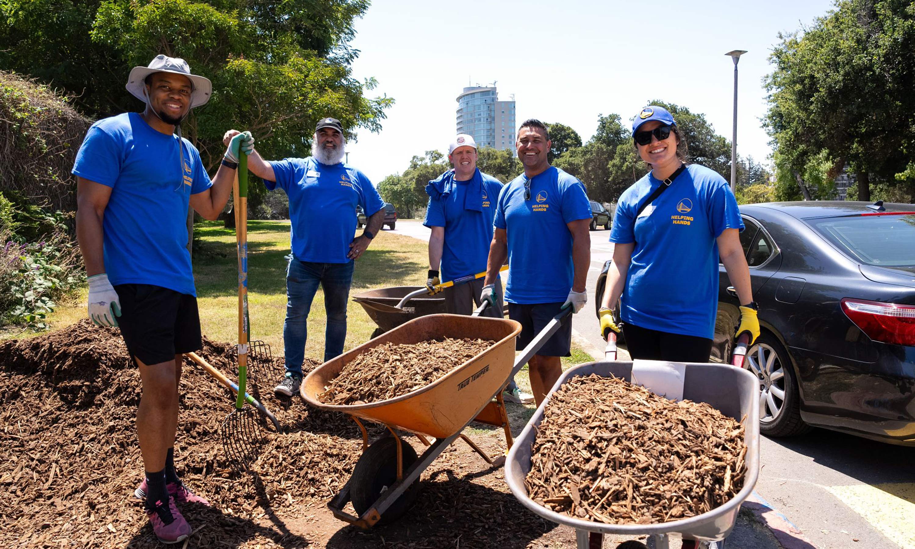 Five warriors employees in volunteer shirts using wheelbarrows to move mulch in a garden.