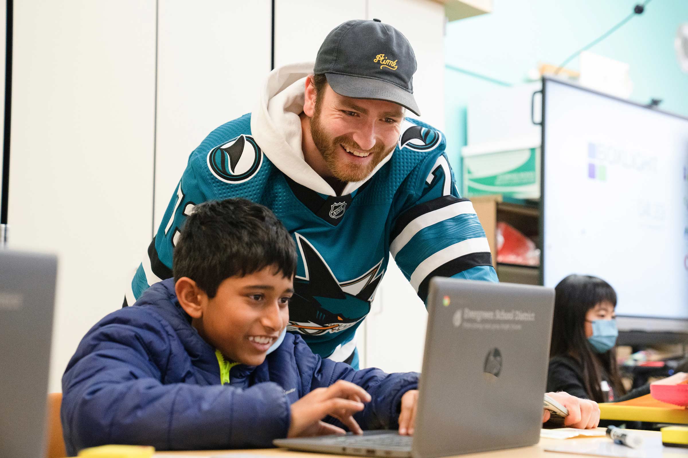 A Shark player in his teal jersey looking over the shoulder of a teenager while he's working on a laptop computer in a classroom.