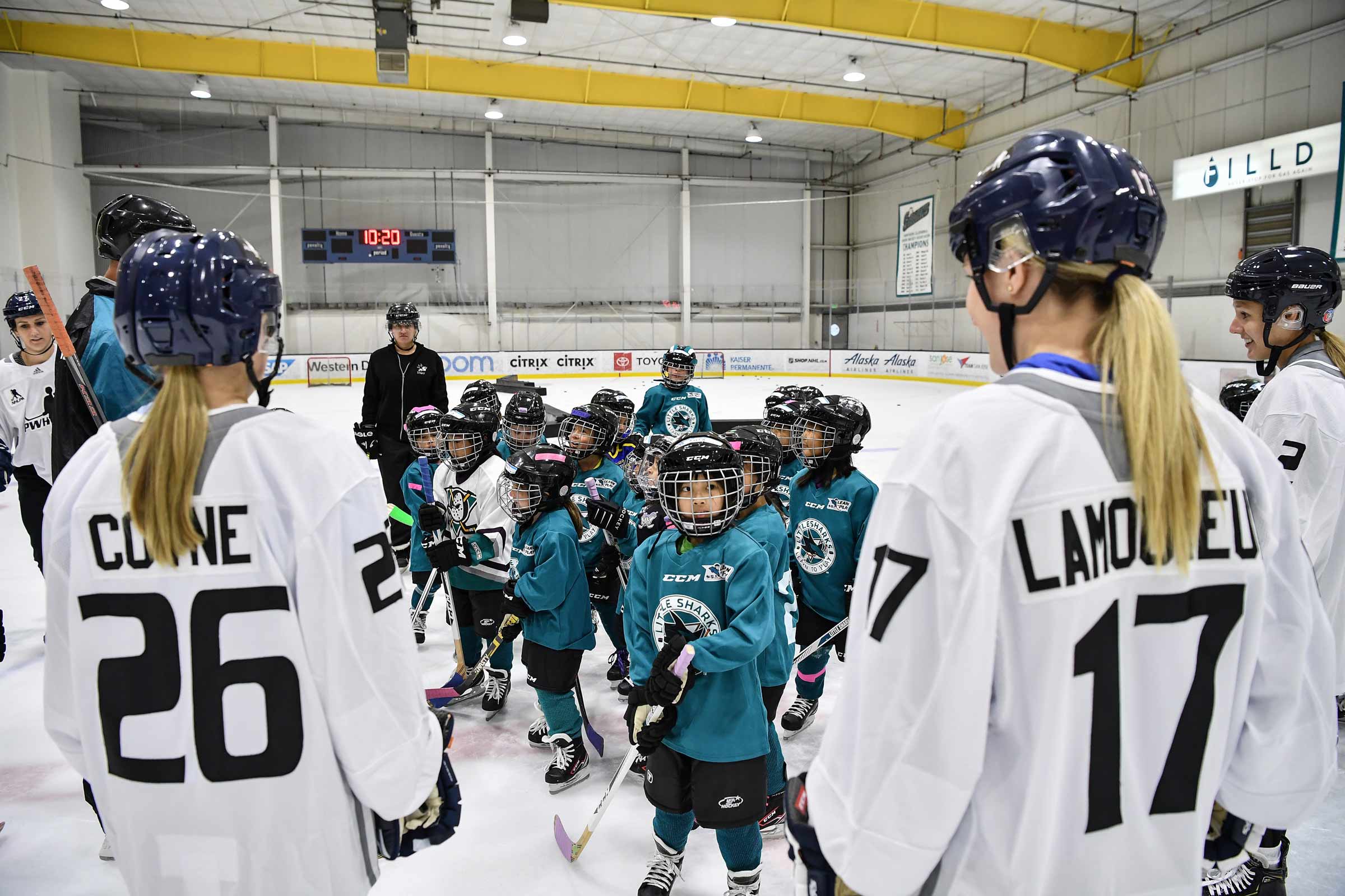 Kids wearing Little Sharks jerseys who are a part of the kid's league on the ice in between play surrounded by women hockey instructors