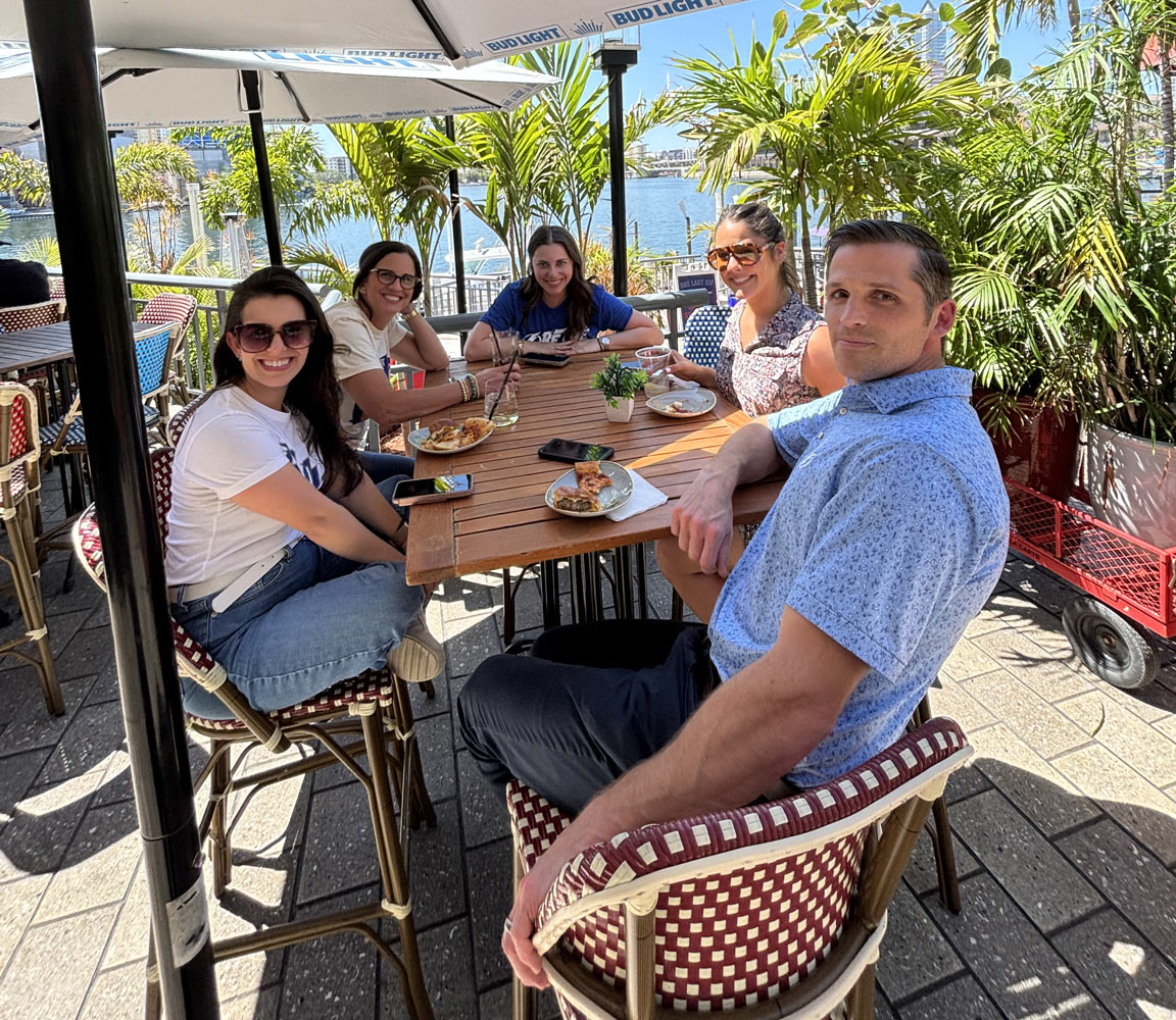 Four adults dining outside at a restaurant with palm trees and the bay in the background.