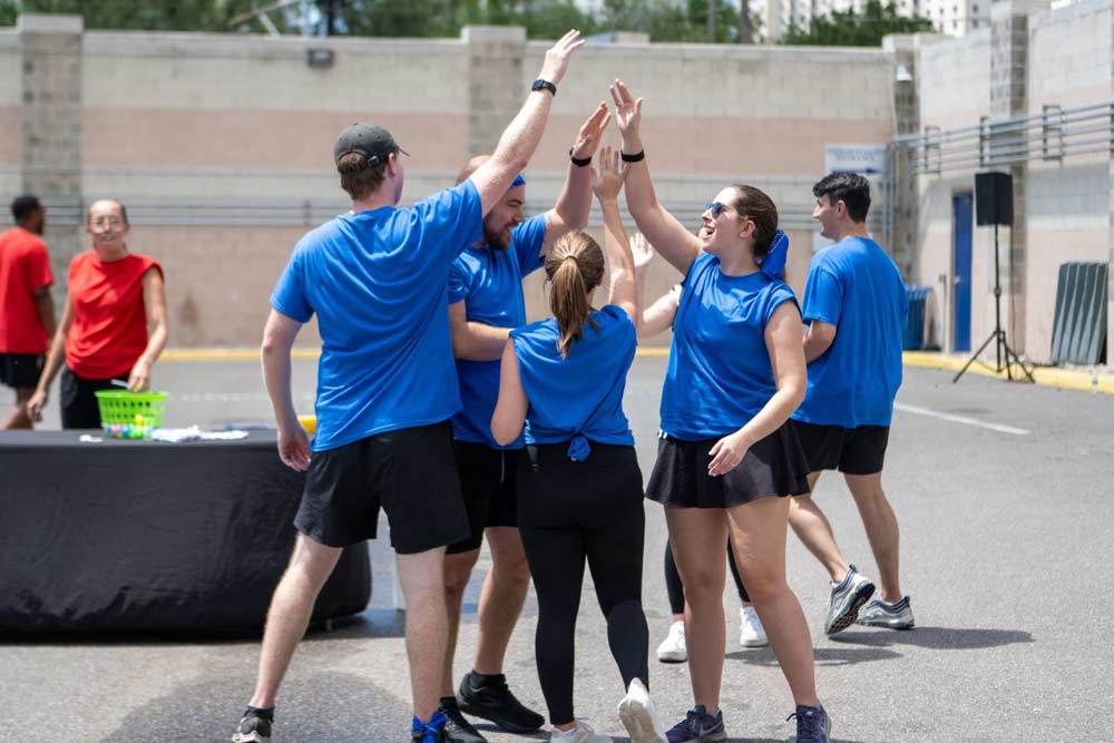 5 young adults high fiving each other outside in matching bright blue t-shirts