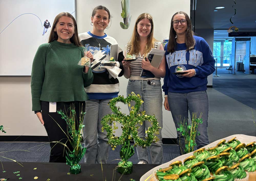 Four women in an office holding cupcakes with green frosting in front of a table with more cupcakes and St. Patrick's Day decorations