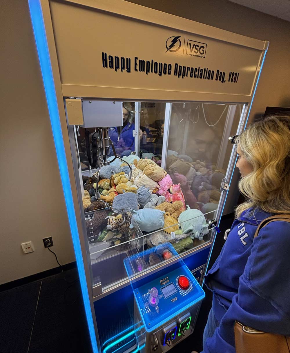 A woman playing a claw machine game that has a sign above it that reads 'Happy Employee Appreciation Day, VSG!'.