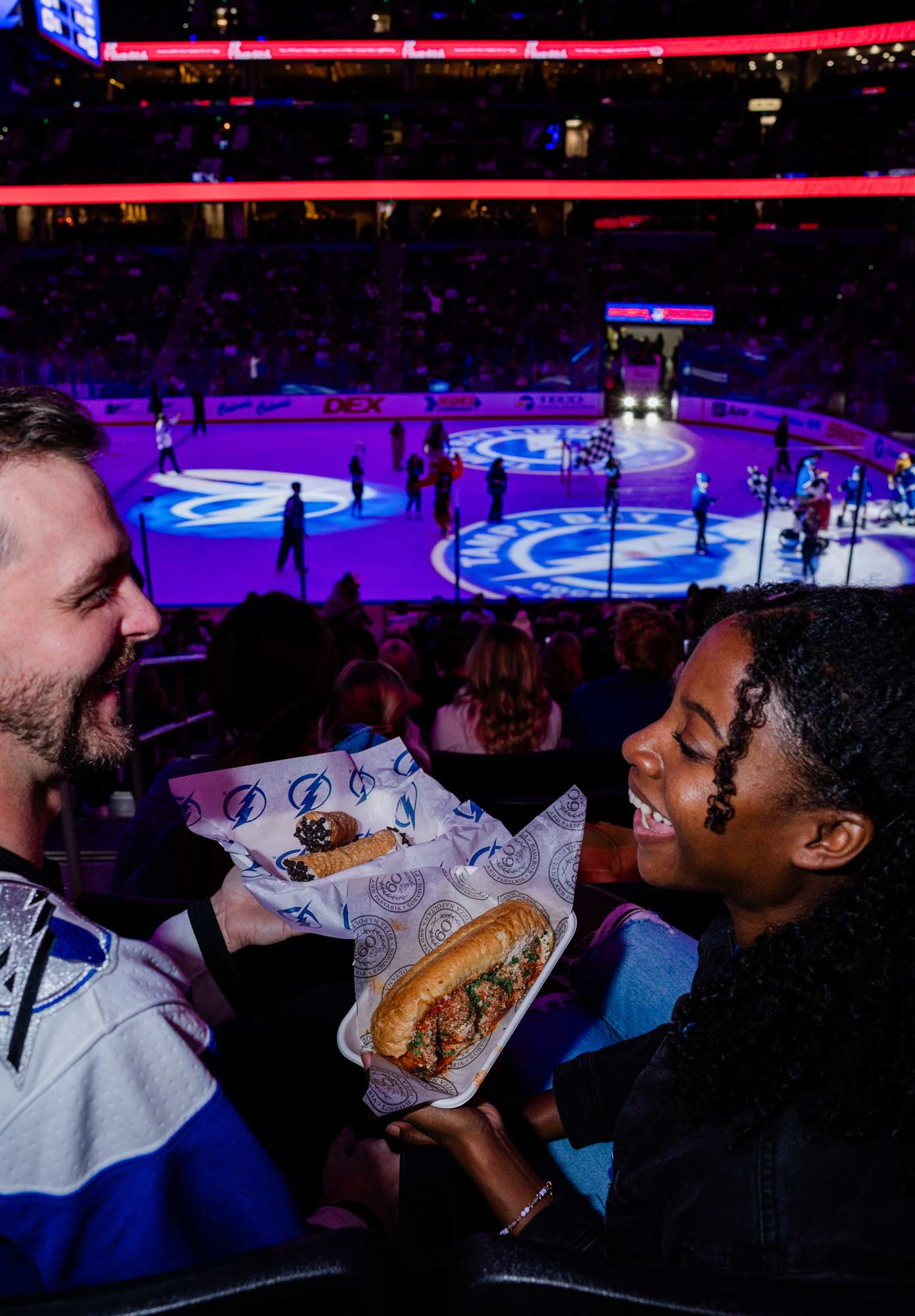 A man and woman sitting in the stands of a hockey game eating meatball subs and cannoli.